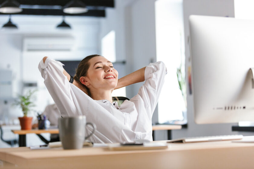 Photo of a smiling woman with arms behind her head, reclining in a chair at a work desk.